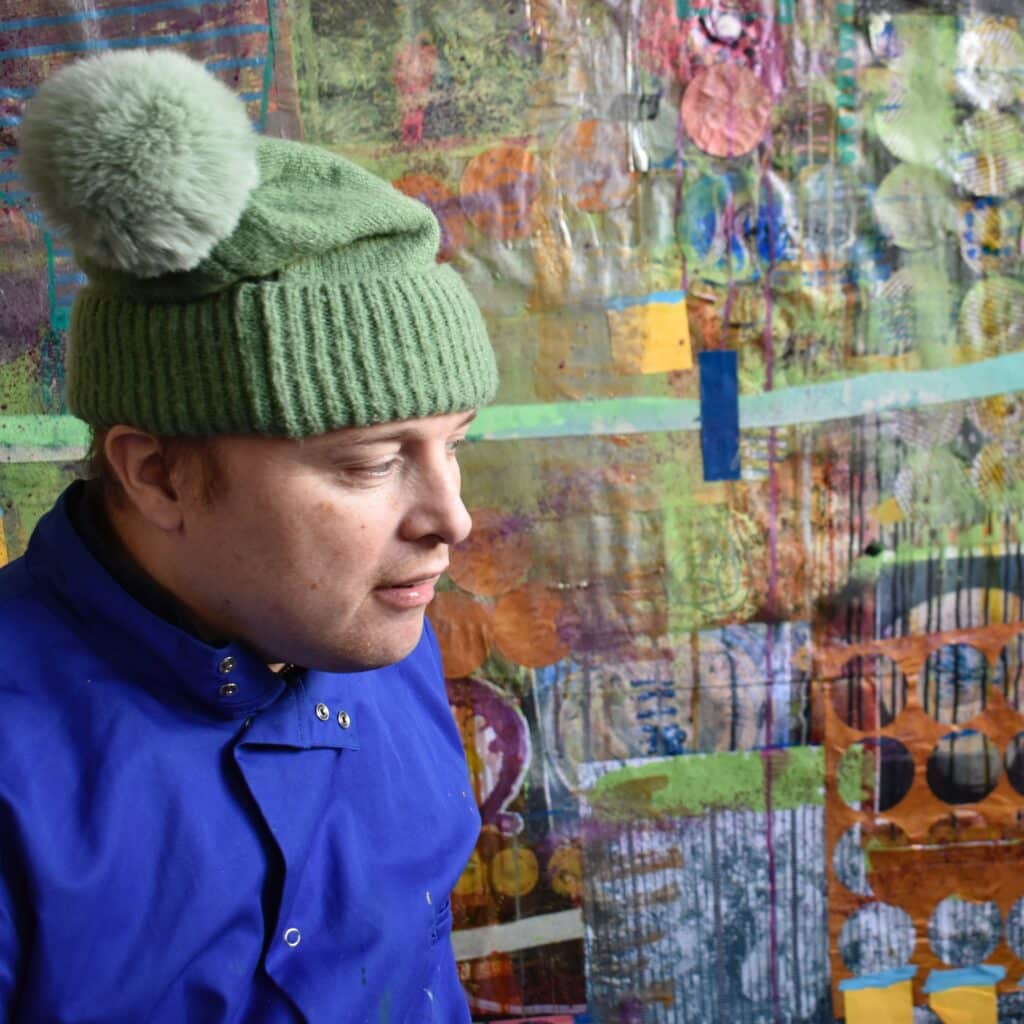 Sean Ormonde stands in front of his work. A large collage. Sean is wearing a green woollen hat with a bobble on top and blue overalls.