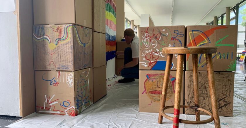 Stacked cardboard boxes are placed in the studio as a man marks them with bright colours and patterns.