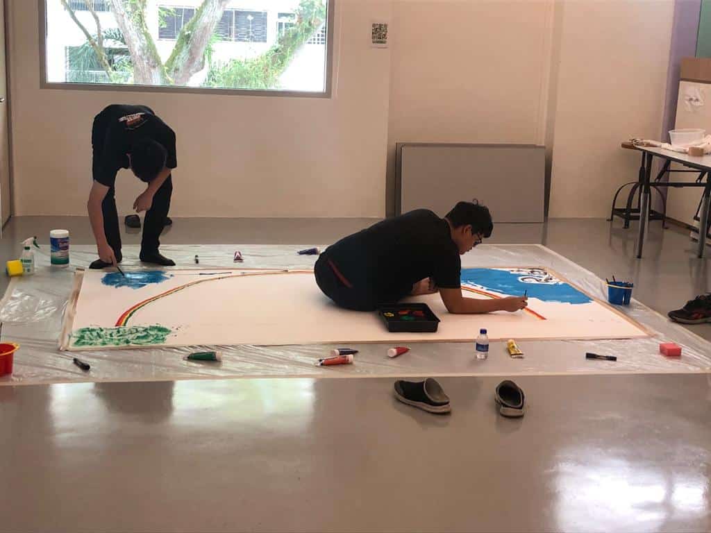 Two people paint on a large piece of paper covering the floor. One lies on top of the paper while the other stands and bends down to make their mark.