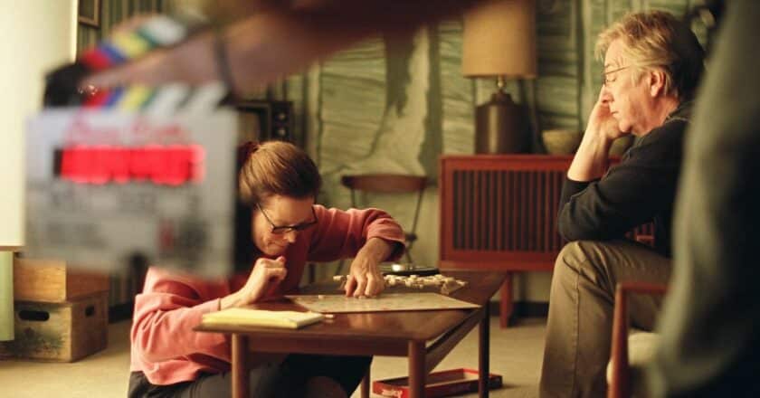 A still from the film Snow Cake released in 2006. in the background a woman sits crossed legged on the floor drawing on paper. A man sits on a chair in front of her watching intently. in the foreground a clapper board is being held up.