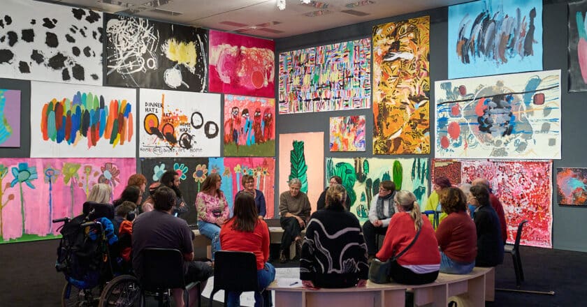 A circle of people sit talking in an exhibition space surrounded by large scale paintings.