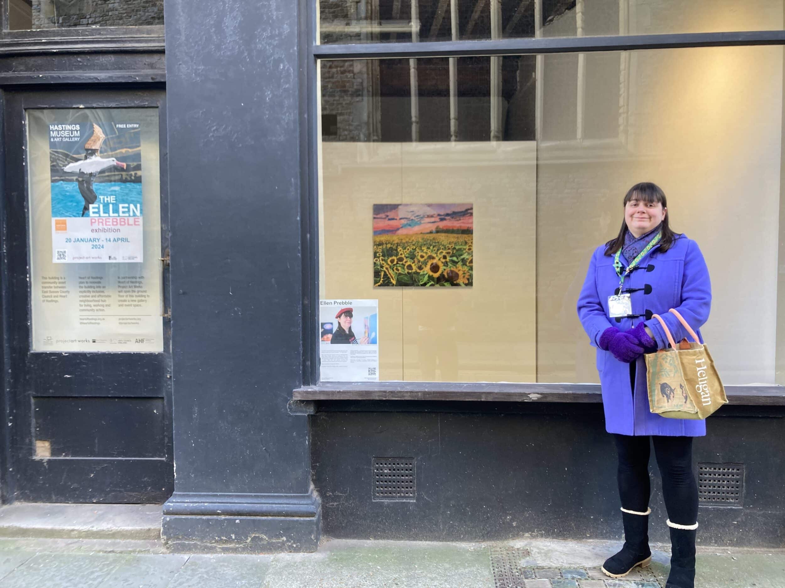 Ellen stands in front of the Untitled Gallery window. In the window you can see her Sunflower Fields painting and a poster for her exhibition.