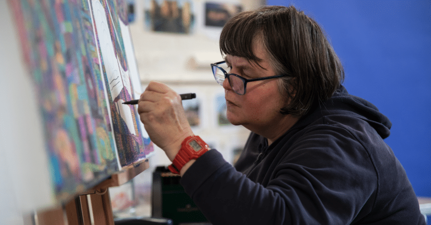 Michelle Roberts in working on a large canvas in the studio. Michelle is using a felt pen on the canvas where you can see some intricate drawing. Michelle has bobbed hair, is wearing a red watch, a navy hoodie and glasses.