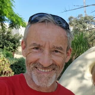 Nick Ewbank smiles at the camera. He is wearing a red t-shirt and has sungalsses on his head. There is a lovely blue sky behind him.