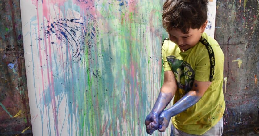 Oliver is a small child wearing a bright yellow t-shirt. They are standing in front of a a wall covered in bright, spattered paint. Oliver looks delighted. They have paint all over their hands and up their forearms.