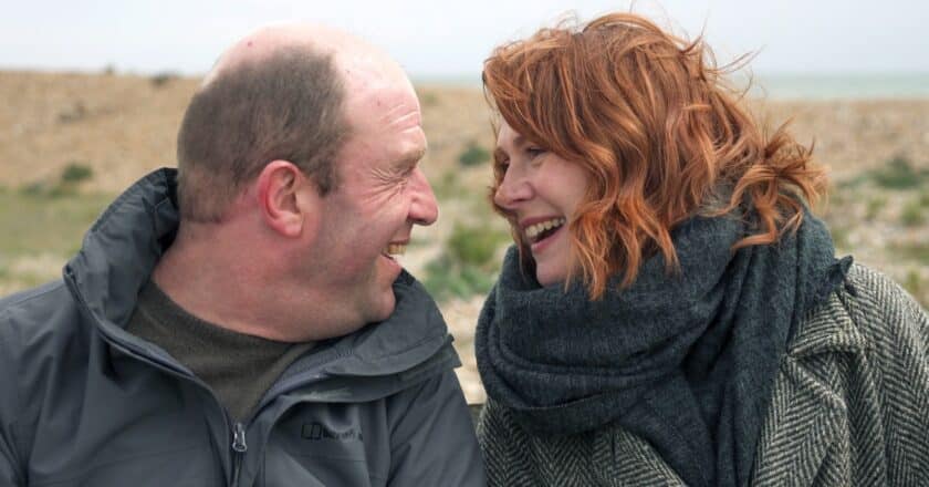 Paul and Ruby Colley look at each other smiling. They are sat outside and are wrapped in warm coats and scarfs. They sit closely together. Ruby has red hair which is flying about in the wind. The landscape behind them looks to be a pebbled beach.