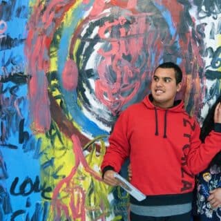 Siddharth and his Mum Susmitha stand in front of Sid's art work. A huge painting on the wall of Phoenix Art Space. Sid has his had resting on his Mum's face. both look happy.