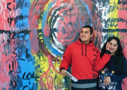 Siddharth and his Mum Susmitha stand in front of Sid's art work. A huge painting on the wall of Phoenix Art Space. Sid has his had resting on his Mum's face. both look happy.