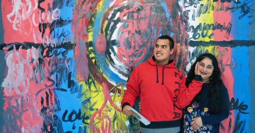 Siddharth and his Mum Susmitha stand in front of Sid's art work. A huge painting on the wall of Phoenix Art Space. Sid has his had resting on his Mum's face. both look happy.