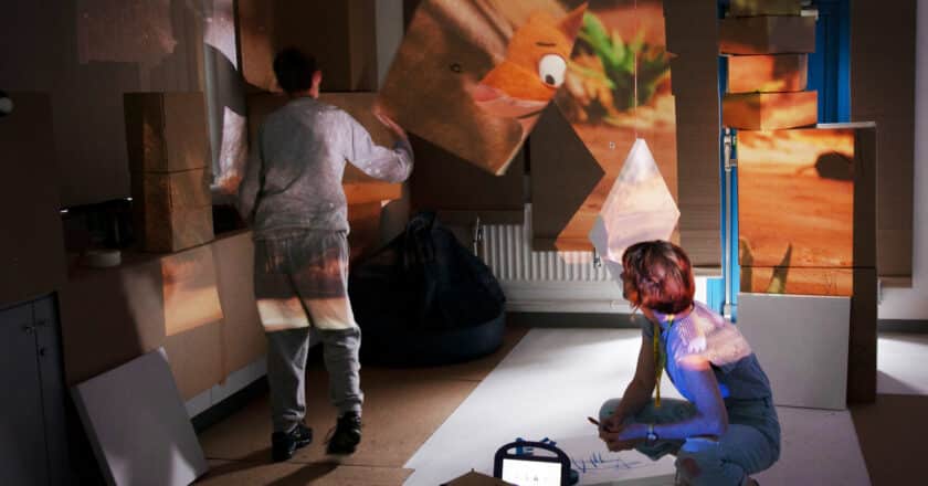 A dark room is filled with cardboard boxes. Projected onto the boxes there is a disjointed image that looks like an animation being played across the box surfaces. It's hard to make out what the image if. An adult stands looking animated in the space. Their hands are up and they are watching the projection. Kneeling away from the standing figure Patricia watches the person. On the floor lays a large piece of white paper with some marks made on it.