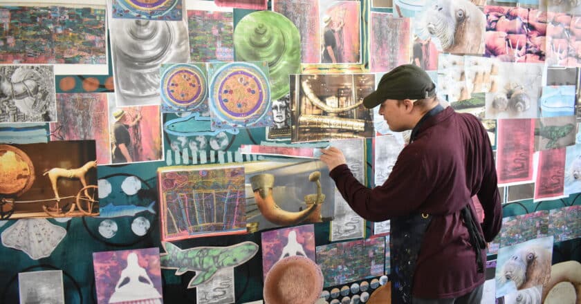 Sean is an artist and is working on a large scale collage. Sean wears a baseball cap, a Burgundy jumper and an apron. He stands at the wall where a large piece of paper is covered in A4 print outs of brightly coloured shapes and patterns.