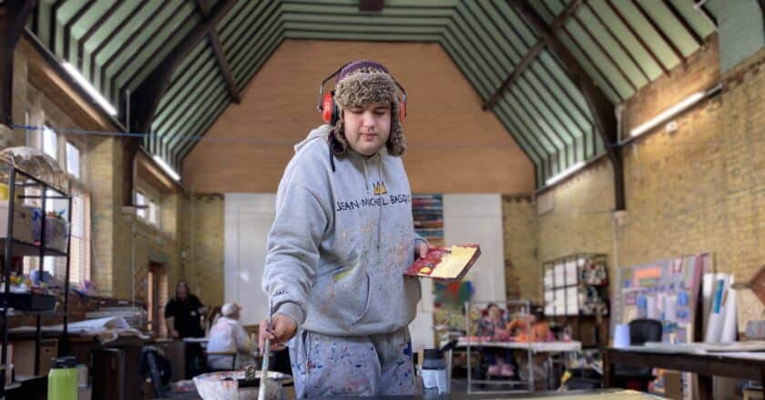 Johnny Pell paints in the large hall of Trinity Hall at Project Art Works. Johnny wears grey tracksuit, a fluffy hat and ear defenders. Behind Jpnny, out of focus, you can see other artists working together.