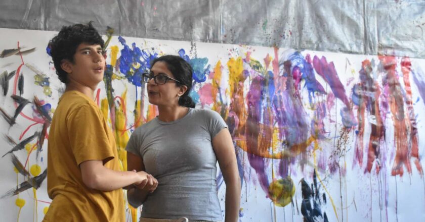 Raffy stands with his Mum in front of a large piece of white paper on the wall. The paper is covered in large strokes of paint moving vertically down. Raffy looks animated and is looking at the camera. His Mum is holding his hand and looking at him. Raffy is tall with a yellow t-shirt and black joggers. His Mum wears a grey T-shirt and joggers and wears her dark hair back in a pony tail.