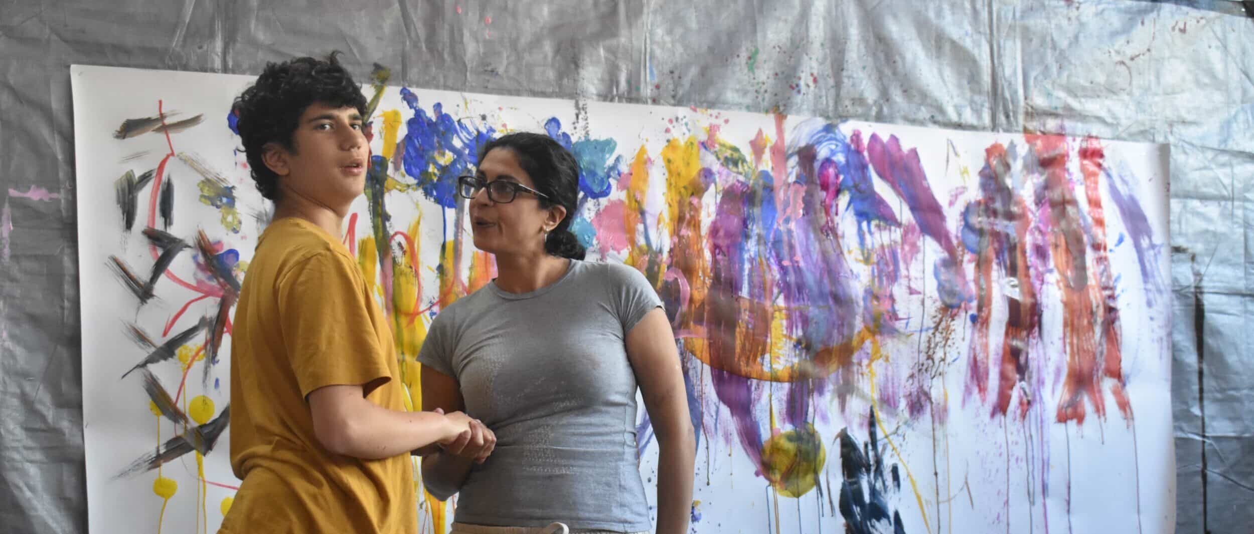 Raffy stands with his Mum in front of a large piece of white paper on the wall. The paper is covered in large strokes of paint moving vertically down. Raffy looks animated and is looking at the camera. His Mum is holding his hand and looking at him. Raffy is tall with a yellow t-shirt and black joggers. His Mum wears a grey T-shirt and joggers and wears her dark hair back in a pony tail.