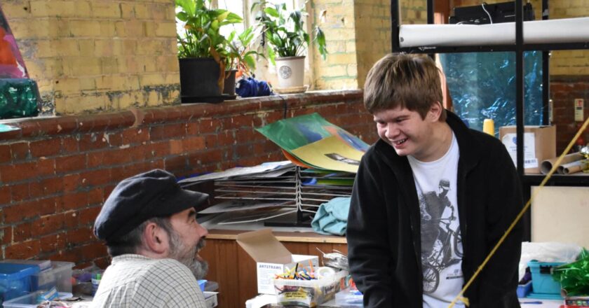 Mark, an artist facilitator, smiles up at a young person called Flinn, who is smiling back. They are in a sunlit room with art works drying behind them and pots full of felt tips.