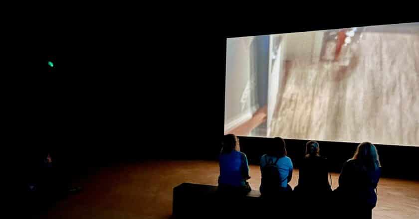 In a dark room a large screen is playing a film. The film itself is a blur and hard to make out what is being shown. In front of the screen four people sit on a bench with their backs to the camera watching the film.