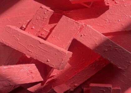 Rectangle blocks of wood are lying on top of each other all covered in pink paint. The paint looks thickly applied with large drops coming off in places. In the photograph shadows are cast across the image adding to the 3d nature of the piece.