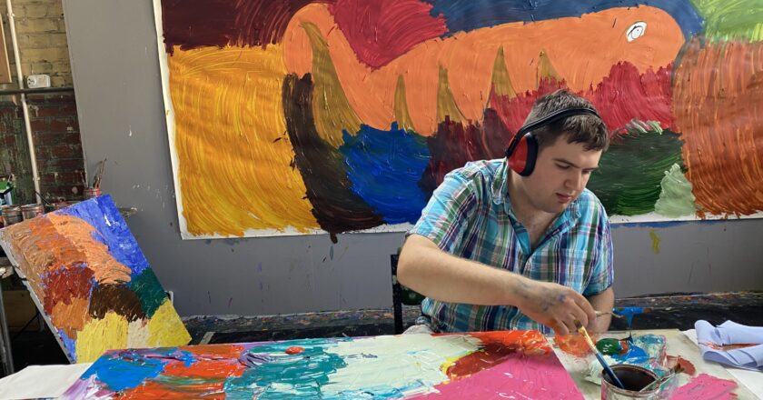 Johnny Pell works in the Project Art Works studio. Johnny is sat at a table with a large canvas in front of him. Johnny is wearing red noise cancelling headphones.
