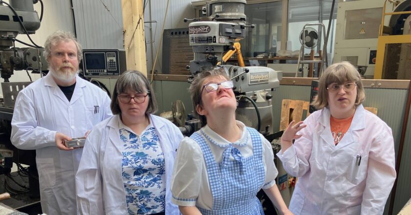 A promotional photo from the film The Memory Blocks, by Andrew Kötting. Neville Jermyn, Michelle Roberts and Charlotte Stephens stand in a row in white lab coats. Eden Kötting wears a blue and while gingham dress, like Dorothy from the Wizard of Oz, and holds onto a stroller.